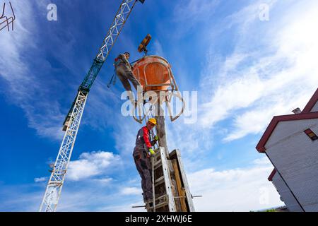 Bauarbeiter entladen konische Metalleimer, gießen Beton in eine hohe vertikale Form für die Säulen des Gebäudes und verwenden einen Kran, während er den Kegel t trägt Stockfoto