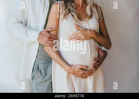 Unerkennbarer Mann und schwangere Frau umarmen den Bauch, während sie in der Nähe der weißen Wand stehen. Stockfoto