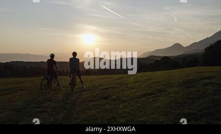 Zwei Silhouetten von Straßenradfahrern, die eine Fahrt mit einem hohen Gruß feiern, bei Sonnenuntergang mit fantastischem Blick auf die Berge, Panoramaaufnahme aus der Luft. Stockfoto