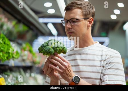 Mann mit Brille und gestreiftem Hemd untersucht Brokkoli-Kopf im Lebensmittelgeschäft. Frisches Gemüse im Supermarkt kaufen. Stockfoto