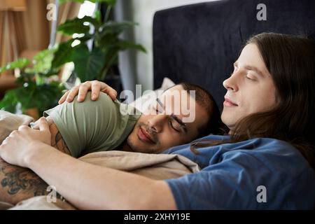 Ein junges schwules Paar ruht friedlich in ihrem Schlafzimmer, ein Mann schläft, während der andere seinen Kopf auf der Brust seiner Partnerin auflegt. Stockfoto