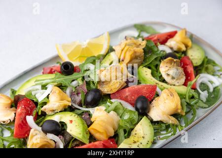 Warmer Salat mit Raps (Schnecken), Tomaten, Avocado, Rucola, schwarzen Oliven, Zwiebeln und Olivenöl. Traditioneller Salat mit Meeresfrüchten. Nahaufnahme, selektiver Fokus. Stockfoto