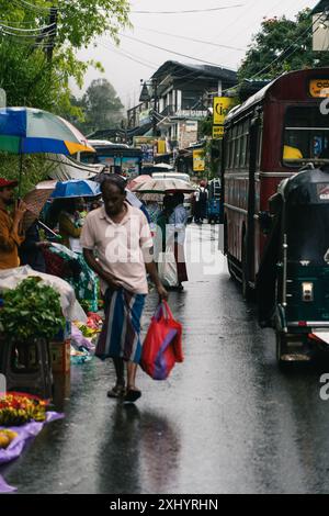 Regnerische Straßenszene mit Leuten, die Regenschirme und Einkaufstaschen auf einer nassen Straße in Ella, Sri Lanka, tragen. Stockfoto