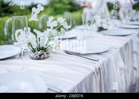 Der festliche Tisch mit weißer Tischdecke ist mit Blumen dekoriert. Leere Teller und Gläser warten auf die Gäste. Ein Festessen im Garten. A Stockfoto