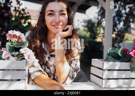 Eine junge Frau mit lockigen Haaren sitzt an einem Tisch auf der Veranda und lächelt. Ein attraktives Mädchen genießt Blumen im Garten. Eine charmante Frau ist ich Stockfoto