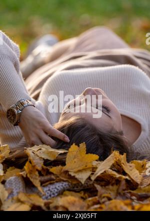 Herbstspaziergang. Weibliches Porträt. Ein glückliches Mädchen liegt mit geschlossenen Augen auf dem Boden in einem Park zwischen gelben Blättern; Seitenansicht, Rückansicht Stockfoto