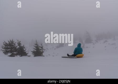 Vormittag aus verschneiten Bergen. Eine Frau sitzt auf Touristenmatratzen und schaut in den Wald. Nebel Winter Stockfoto