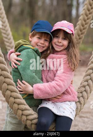 Das Mädchen umarmt ihren Bruder zärtlich. Kinder sitzen auf einer Schaukel im Stadtpark und lachen. Bruder und Schwester, Kinder, Familie, Freundschaft Stockfoto