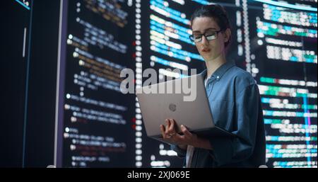 Porträt einer Frau, die eine Software und Codierung erstellt, umgeben von großen Bildschirmen, die Zeilen der Programmiersprache anzeigen. Programmiererin, die in einem Überwachungsraum arbeitet. Futuristisches Konzept Stockfoto