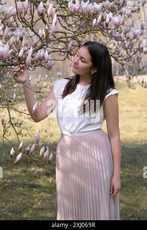 Frühlingsporträt eines jungen, dunkelhaarigen Mädchens in blühender Magnolie. Teenager-Modell in der Nähe eines Baumes mit Rosenblüten Stockfoto