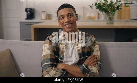 Lächelnder junger Erwachsener afroamerikanischer Mann sitzt entspannend auf der Couch Wohnung birassischer Hausbesitzer ethnischer männlicher Mann lächelnd die Hände kreuzend in die Kamera schauend Stockfoto