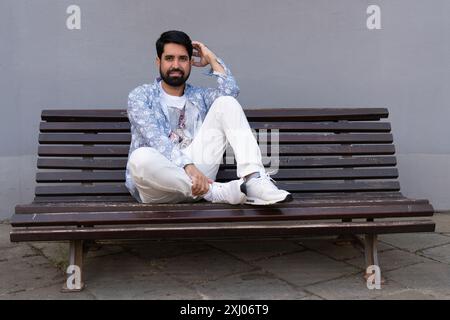 Ein moderner, stilvoller pakistanischer Mann mit dunklem Bart, in hellen Kleidern, sitzt auf einer Holzbank Stockfoto