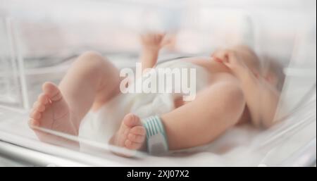 Niedliches kleines kaukasisches Neugeborenes, das im Bassinet in einem Entbindungskrankenhaus liegt. Porträt eines winzigen, verspielten und energiegeladenen Kindes mit einem Namensschild auf dem Bein. Gesundheit, Schwangerschaft und Mutterschaft Konzept Stockfoto