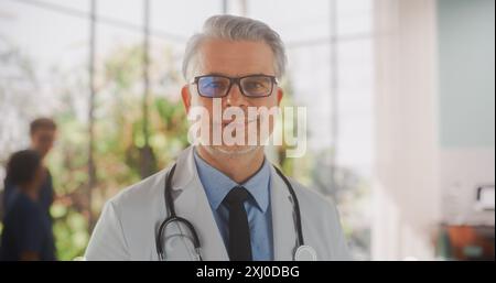 Porträt eines Mediziners mittleren Alters, der posiert, lächelt und in die Kamera schaut. Kaukasischer Klinikarzt mit Brille und weißem Mantel in der Krankenhauspraxis Stockfoto
