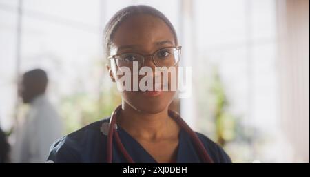 Porträt einer schönen afrikanischen Ärztin, die die Kamera ansieht und lächelt. Junge Gesundheitsfachkraft rettet Leben, arbeitet im modernen Krankenhaus. Attraktive schwarze Krankenschwester in stilvoller Brille Stockfoto