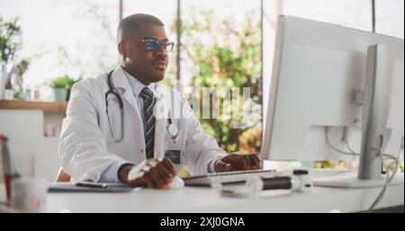 Junger afrikanischer männlicher Arzt mit weißem Mantel, der in seinem Büro am PC arbeitet. Handsome Black Medical Care Professional arbeitet mit Testergebnissen, Patientenbehandlungsplanung Stockfoto