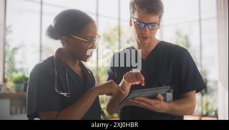 Junge positive männliche und weibliche Krankenschwester stehen in einem Flur eines modernen öffentlichen Gesundheitszentrums. Afrikanische Frau führt ein Gespräch mit einem Kollegen und diskutiert die Behandlung auf einem Tablet-Computer Stockfoto