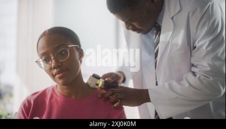 Afroamerikanischer Dermatologe mit einem Dermatoskop, um besorgniserregende krebserregende Gewebe auf der Haut einer schönen jungen Frau während eines Health Check Besuchs in einer Klinik zu identifizieren. Schwarzer Arzt im Krankenhaus Stockfoto