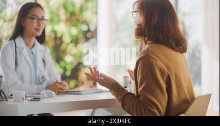 Die Allgemeinärztin führt ein Gespräch mit einer älteren Patientin und spricht mit ihr während der Konsultation in einer Gesundheitsklinik. Arzt im Labormantel, der hinter einem Schreibtisch im Krankenhausbüro sitzt Stockfoto