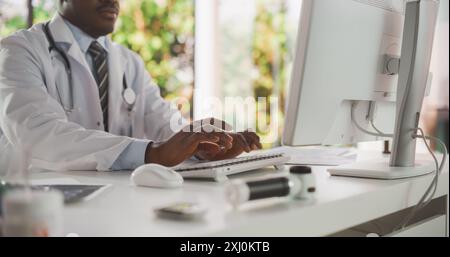 Nahaufnahme eines afrikanischen männlichen Arztes in White Coat, der am Computer an seinem Schreibtisch im Büro arbeitet. Black Medical Care Professional mit Tastatur, Arbeiten mit Patiententerminen Stockfoto
