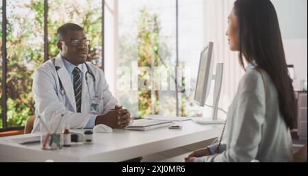 Handsome Young African Doctor führt ein Gespräch mit einer multiethnischen asiatischen Patientin und spricht mit ihr während der Konsultation in einer Gesundheitsklinik. Arzt im Weissmantel im modernen Krankenhaus Stockfoto