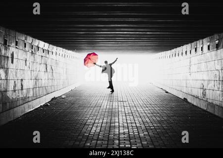 Silhouette eines Mannes mit rotem Regenschirm in einem Tunnel. Schwarzweißbild mit Licht von hinten. Person, die auf die Kamera zuspringt. Stockfoto