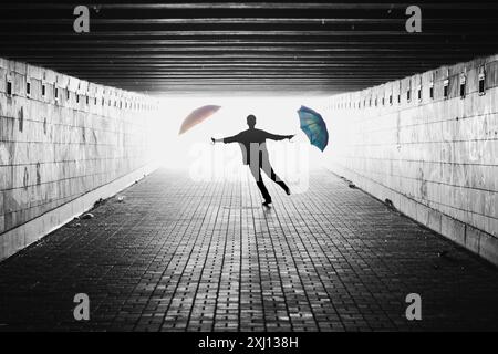 Silhouette eines Mannes mit zwei Regenschirmen in einem Tunnel. Schwarzweißbild mit Licht von hinten. Person, die auf die Kamera zuspringt. Stockfoto