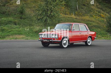 Rote klassische VAZ 2103 Limousine in der Nähe eines grünen Hügels. Drei Viertel Vorderansicht von Lada aus den 1970er Jahren auf einem großen, leeren Parkplatz. Stockfoto