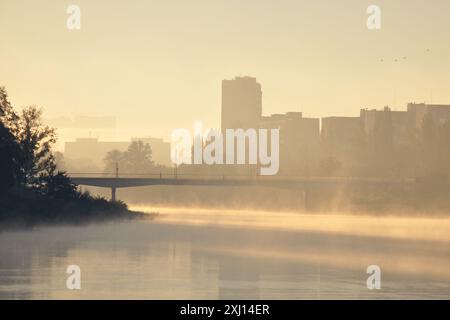 Nebeliger Sonnenaufgang in einer Stadt. Nebel auf dem Wasser des Flusskanals. Fußgängerbrücke und Silhouetten von Gebäuden, die von der Morgensonne hinterleuchtet werden. Stockfoto