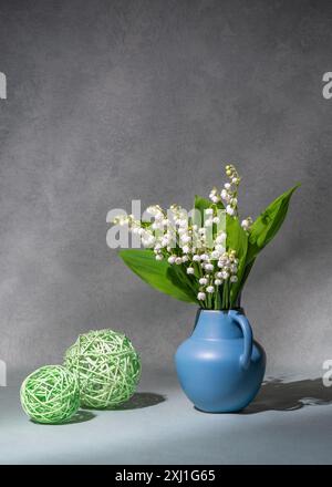 Ein Blumenstrauß aus Frühlingsblumen, Maiglöckchen aus Tal in einer blauen Vase, auf grauem Hintergrund. Es gibt zwei grüne Bälle in der Nähe. Stillleben, Postkarte, Kopie Spac Stockfoto