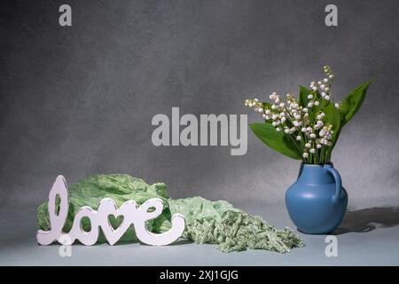 Ein kleiner Strauß weißer Blumen, Maiglöckchen aus dem Tal in einer blauen Vase neben der Aufschrift „Liebe“. Grauer Hintergrund. Platz für Text. Stillleben. Pos. Stockfoto