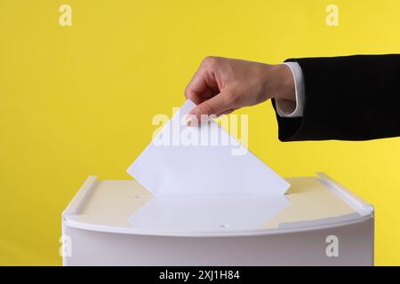 Frau, die ihre Stimme vor gelbem Hintergrund in die Wahlurne setzt, Nahaufnahme Stockfoto