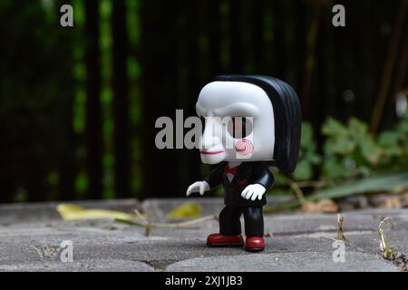 Funko Pop Actionfigur von Billy der Puppe aus dem beliebten Slasher-Horrorfilm SAW. Dunkler Hof, schwarzer Zaun, Schatten, stimmungsvoller grüner Hintergrund. Stockfoto