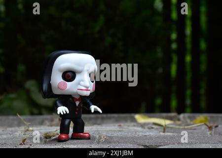 Funko Pop Actionfigur von Billy der Puppe aus dem beliebten Slasher-Horrorfilm SAW. Dunkler Hof, schwarzer Zaun, Schatten, stimmungsvoller grüner Hintergrund. Stockfoto