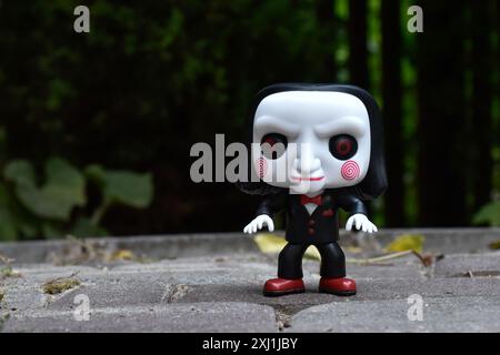 Funko Pop Actionfigur von Billy der Puppe aus dem beliebten Slasher-Horrorfilm SAW. Dunkler Hof, schwarzer Zaun, Schatten, stimmungsvoller grüner Hintergrund. Stockfoto