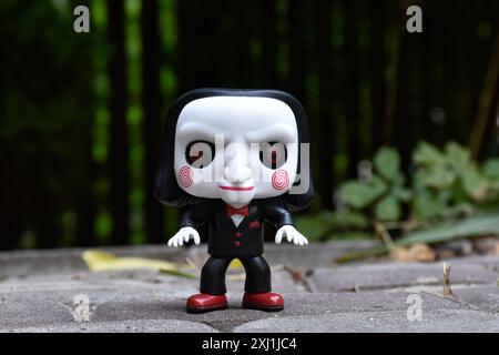 Funko Pop Actionfigur von Billy der Puppe aus dem beliebten Slasher-Horrorfilm SAW. Dunkler Hof, schwarzer Zaun, Schatten, stimmungsvoller grüner Hintergrund. Stockfoto