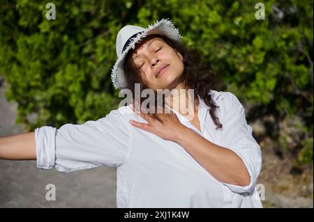 Eine Frau in einem weißen Hemd und Hut genießt einen friedlichen Moment mit geschlossenen Augen und ausgestrecktem Arm in der Natur. Entspannungs- und Ruhekonzept. Stockfoto