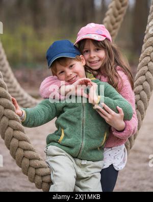 Porträt eines wunderbaren Bruders und einer wunderbaren Schwester. Das Mädchen umarmte den Jungen und faltete ihre Hände zu einem Herz. Liebe in der Familie. Kinder spielen auf einer Seilschaukel Stockfoto