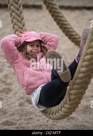 Ein fröhliches, glückliches Mädchen von 7 Jahren spielt glücklich in der Natur in einem Stadtpark. Auf einem dicken Seil liegend, auf einem Karussell. Kind ist eine Kappe Stockfoto