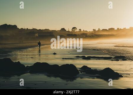 Silhouette eines Surfers, der am Ufer des Strandes mit einem wunderschönen Sonnenuntergang spaziert. Patos Beach in Nigran. Galicien - Spanien Stockfoto