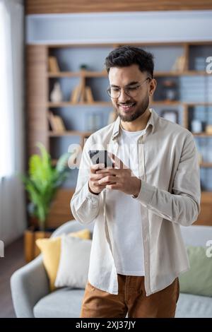 Vertikale Aufnahme eines jungen Indianers zu Hause, der in einem Zimmer steht und lächelt, während er ein Handy benutzt. Stockfoto