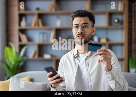 Nahaufnahme eines aufgebrachten jungen Indianers zu Hause, der Kreditkarte und Telefon hält und besorgt in die Kamera schaut. Stockfoto