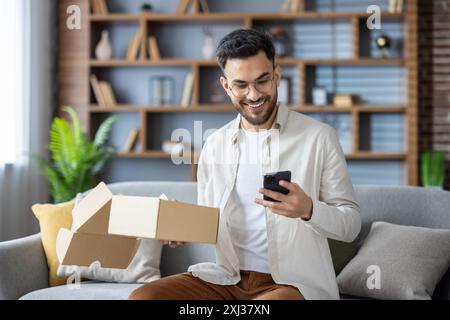 Lächelnder Mann, der auf der Couch sitzt und das Paket auspackt, während er das Smartphone überprüft. Modernes Wohnzimmer mit gemütlichem Ambiente, Topfpflanzen und Bücherregalen im Hintergrund. Konzept des Online-Einkaufs und der Lieferung. Stockfoto