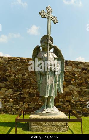 Astorga, Spanien – 4. Juni 2023: Vor einer Steinmauer steht eine bronzene Engelsstatue, die ein Kreuz in die Höhe hält. Die Statue ist von grünem Gras umgeben. Stockfoto