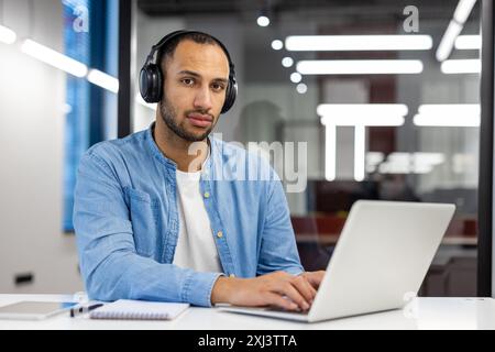 Porträt eines ernsthaften hispanischen Mannes, der im Büro mit Kopfhörern an einem Schreibtisch sitzt, auf einem Laptop arbeitet und schreibt und auf die Kamera blickt. Stockfoto
