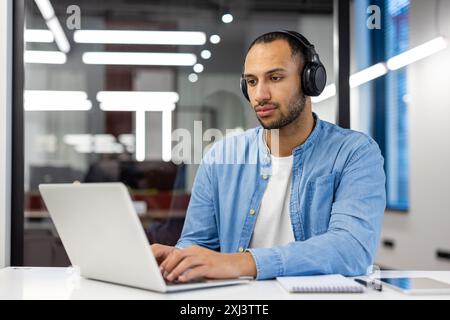 Konzentrierter junger muslimischer Mann, der in modernen Büros an einem Laptop arbeitet, am Schreibtisch sitzt und Kopfhörer trägt. Stockfoto