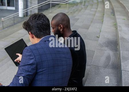 Zwei multikulturelle Geschäftsleute, ein afrikanischer und der andere hispanischer, in Anzügen gekleidet, werden von hinten gesehen, während sie an einem Laptop arbeiten, während sie sitzen Stockfoto