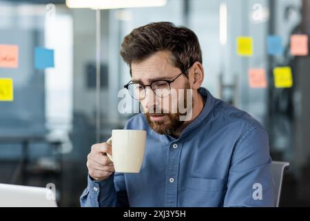 Ein müder Geschäftsmann sitzt im Büro und hält eine Kaffeetasse, trägt eine Brille und blickt auf den Laptop-Bildschirm. Das Konzept von Stress, Ermüdung und Arbeitsdruck im modernen Geschäftsumfeld Stockfoto