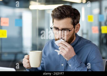 Nahaufnahme eines kranken jungen männlichen Büroangestellten, der an seinem Arbeitsplatz sitzt, eine Tasse heißes Getränk und Medikamente in der Hand hält und sich mit einer Serviette von einer laufenden Nase abwischt. Stockfoto