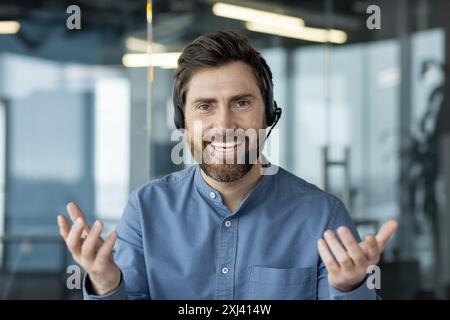 Porträt eines jungen lächelnden Mannes in einem blauen Hemd und mit Bart, der ein Headset trägt, vor der Kamera im Büro sitzt und einen Videoanruf führt, mit den Händen gestikuliert. Stockfoto